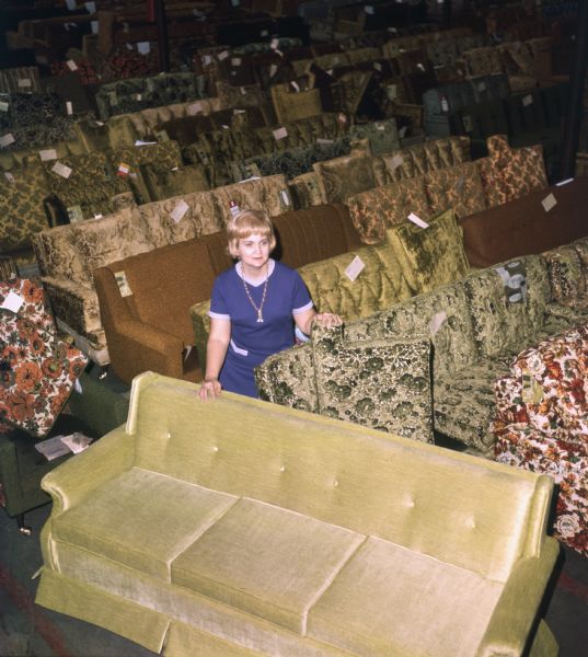 Elevated view of a blonde haired woman in a warehouse full of sofas or couches at Mastercraft Furniture. She is sitting on the arm of one of the sofas and is wearing a blue dress with white trim and a necklace.