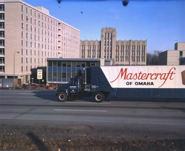 View from side of road of a diesel powered International Harvester Fleetstar 2000-D furniture delivery truck. The cab has an air shield on top to reduce wind drag. The side of the trailer says: "Mastercraft of Omaha," and the truck is owned by Mastercraft Furniture Corporation of Omaha, Nebraska. A speed limit sign is on one side of the road and several buildings, as well as a few parked vehicles, can be seen in the background.