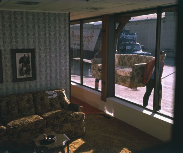 View through window of two men delivering a couch or sofa from Mastercraft Furniture Corporation of Omaha, Nebraska. Inside the room is a couch with tags on it, carpeting, wallpaper with a decorative pattern, a small table with an ashtray, and an illustration hanging on the wall. One of the delivery men is wearing a red jacket, and their delivery vehicle, which can be seen in the background, is a diesel powered International Harvester Fleetstar 2000-D with an air shield on top to reduce wind drag. "Mastercraft of Omaha" can be seen on the side of the trailer.