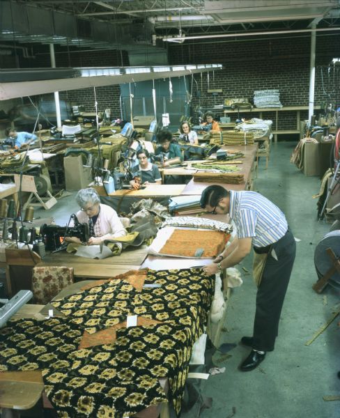 Elevated view of room in which several women sitting and sewing machines are sewing pieces of material for couches or sofas. A man is standing in the aisle cutting material. The original caption reads: "Craftsmanship is evidenced throughout Mastercraft plant. As shown in side-by-side views, women sew backs and arm cushions for sofas at machines."