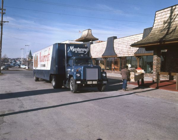 Two men are delivering a couch from Mastercraft Furniture Corporation of Omaha, Nebraska to a furniture store. The men are outside carrying the couch towards the store entrance, and their delivery vehicle is a diesel powered International Harvester Fleetstar 2000-D. "Mastercraft of Omaha" can be seen on the side of the trailer. In the background many parked cars and an American flag can be seen. The original caption reads: "Delivery to Omaha's Paramount Furniture Store (above) illustrates trucks' direct-to-outlet service. To cut fuel costs, firm's linkups use air shields, fitted to top of 2000-D cabs, in concert with vortex stabilizers, which are mounted on vans' fronts. Together they reduce wind 'drag.'"