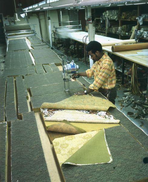 An African-American man, William McQuinn, is cutting material for sofas. He is wearing a striped shirt and standing at a long bench. He is using what appears to be an electric saw with a vertical blade. The original caption reads: "Craftsmanship is evidenced throughout Mastercraft plant. As shown in side-by-side views... William McQuinn cuts, 40 thicknesses at a time, material for sofas."