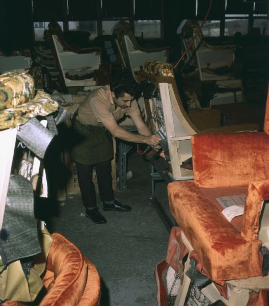 A man is affixing fabric to a chair with a pneumatic stapler. He is leaning forward and is wearing an apron. Piles around him are chairs, pieces of partially covered furniture, and pieces of fabric.