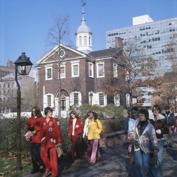 A group of young people walking down a street. They are all wearing coats. A brick building that may be Carpenters' Hall is in the background.