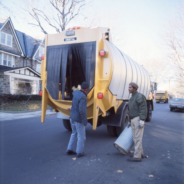 Two workers are placing refuse in a garbage truck. One is an African-American man in a jacket, hat, and gloves and carrying a trash can. The other man is facing away from the camera, and appears to have just thrown something in the back of the truck. A small sign that reads "Orbital" is on the back of the truck. A house, trees, and other and another garbage truck are in the background.