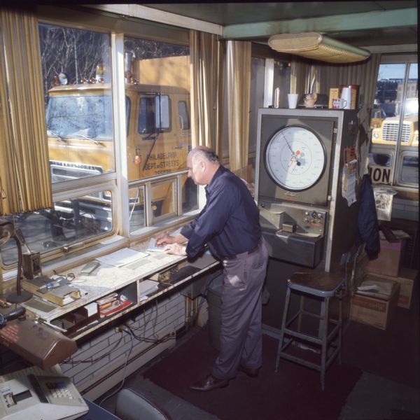 An older man is standing in an office, possibly a weigh station of the Philadelphia Street Department. He is at a bench with papers and various office supplies, and there is a stool behind him. There is also a machine which appears to be a scale, likely for weighing trucks. Through the window can be seen an International Harvester Cargostar model CO-1950.
