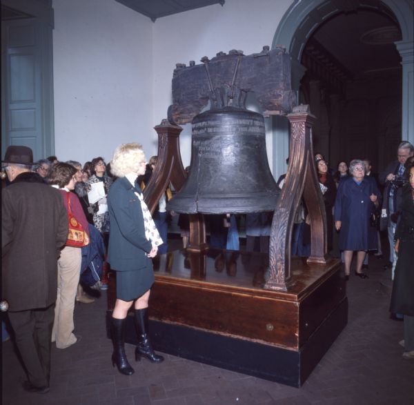 A blonde woman in black boots is standing beside the Liberty Bell. Behind her is a crowd of people.