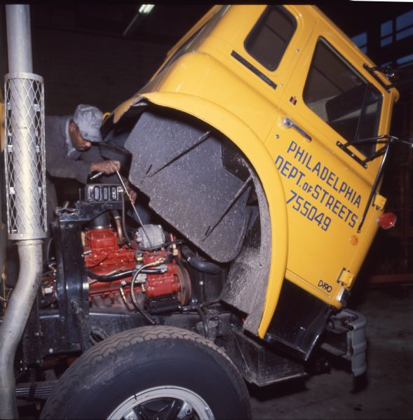 An African-American man is working on a truck engine. He is wearing a hat and overalls, and is using what appears to be a ratchet and extension. The truck is an International Harvester Cargostar model CO-1950, and the cab has been tilted forward to provide access to the engine. The door of the cab has a sign thats: "Philadelphia Department of Streets 755049 D-190."