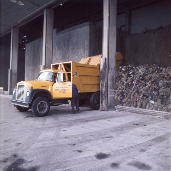 A garbage truck is backed up and emptying refuse into what is likely an incinerator. A man is standing at the open driver's door, and another man is sitting in the cab. The doors a sign that reads: "Philadelphia Department of Streets 755140." The truck is an International Harvester Cargostar model CO-1950.