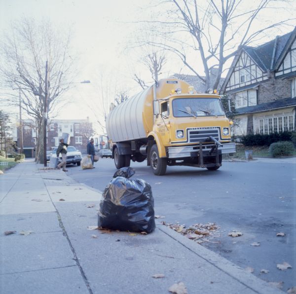 View down sidewalk of two African-American sanitation workers carrying cans and sacks of trash to a garbage truck, is stopped in a residential neighborhood with houses, trees, sidewalks, and parked automobiles. The truck is an International Harvester Cargostar model CO-1950 with an Orbital or Orbie brand body.