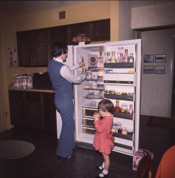 A woman and her three-year-old daughter are getting glasses of milk from a container in a refrigerator. The mother is dressed in a pant suit, while her daughter is in a red dress with white trim, black shoes, and white socks. Kitchen drawers and cabinets can be seen in the room to the woman's left. The photo caption reads: "Mrs. Richard Boland, who is shown with her daughter Mary Clayton, 3, is a regular customer of the routeman, Beryl Fentress."