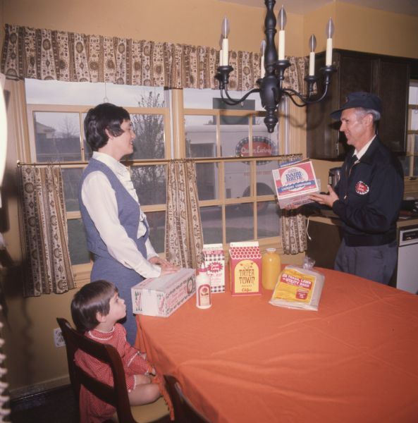 A woman and her daughter are pictured with a delivery man. They are gathered around a table, with the two adults standing and the child sitting. The woman wears a pantsuit, the child wears a dress, and the delivery man wears a uniform. The table has several products on it, including "Tater Town" Potato Chips. The delivery man is holding a box of Dairi-Brite Laundry Compound. A chandelier is hanging from the ceiling, and through the window an International Harvester Loadstar model 1600 with its side door open can be seen. The photo caption reads: "Mrs. Richard Boland, who is shown with her daughter Mary Clayton, 3, is a regular customer of the routeman, Beryl Fentress."