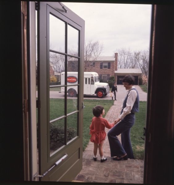 View from inside front door of a mother and her three-year-old daughter standing on steps while a delivery man comes down the walkway.  The mother is dressed in a pantsuit, while her daughter is in a red dress, black shoes, and white socks. The delivery man is in a uniform. An International Harvester Loadstar model 1600 with "Oscar Ewing" written on the side is parked on the street. The photo caption reads, "Mrs. Richard Boland, who is shown with her daughter Mary Clayton, 3, is a regular customer of the routeman, Beryl Fentress."