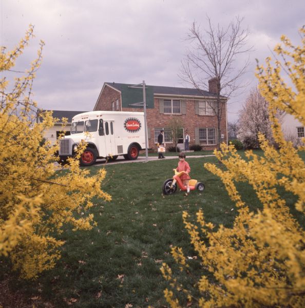 A child (Mary Clayton) is playing on a plastic trike with a large front wheel in a yard. She is wearing a red dress and is standing over the seat of the tricycle. In the background her mother (Mrs. Richard Boland) and a delivery man are standing in front of the house. An International Harvester Loadstar model 1600 with "Oscar Ewing" written on the side is parked in the driveway.
