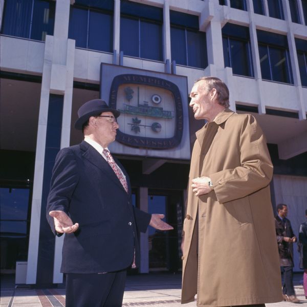 Two men are standing and talking in a plaza in front of a building which has a large sign with the words: "Memphis Tennessee Shelby County" around the seal of the city of Memphis. The seal has depictions of a steamboat, cotton boll, oak tree, and wheel. One man wears a suit, tie and hat, and the other man a coat. An African-American man in a leather coat is in the background. The photo caption reads: "Memphis refuse collection, disposal program is spearheaded by M.C. Stiles who is shown at right (top right) with IH salesman Joe Hill."