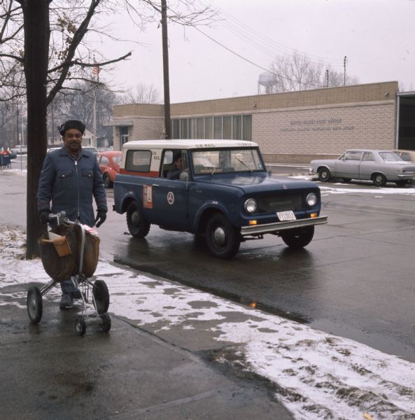 An African-American postal carrier is standing on the sidewalk with mail in a rolling cart. He is wearing a winter postal employee uniform and a hat. Behind him a man is driving a right-hand drive International Harvester Scout, likely a model 800A, with "U.S. Mail" written above the windshield. Across the street is a post office, and behind it in the distance is a water tower.