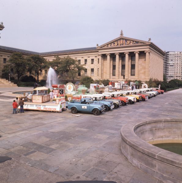 International Harvester Scouts and Travelalls hooked to floats for a Fire Prevention Week parade. The trucks are parked in a line in a plaza near two fountains. In the background is a large stone building with columns. Three children are looking at the closest float, which has the words "[----] Up a Good Recipe for Fire Prevention."