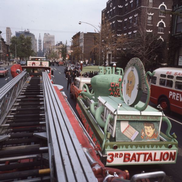 Looking down the length of a fire engine ladder truck during a Fire Prevention Week Parade. On the right is a float with a picture of a child and the words "A child alone fire prone" and "3rd Battalion." The float is being pulled by an International Harvester Scout or Travelall. Firefighters in uniform are further down the street, and in the far background are skyscrapers.