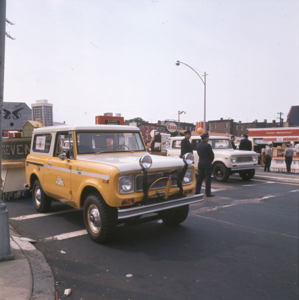International Sno-Star Scout towing a float for Fire Prevention Week. The vehicle is yellow and white with large "fog lamps" mounted on the front. A white International Harvester vehicle painted white is parked alongside. Two uniformed police officers are standing in the street between the two vehicles. On the other side of the street is an Esso service station.