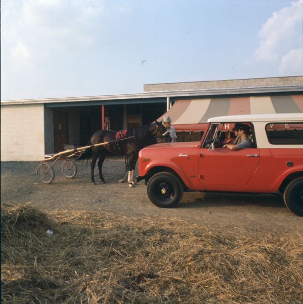 Two police officers are sitting in a red and white International Scout. They are facing a man standing with a horse in front of a stable. The horse is pulling a sulky, likely for harness racing.