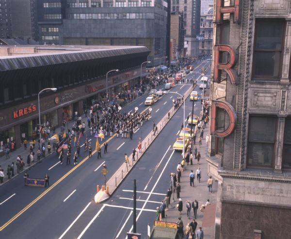 Elevated view of a Fire Safety Prevention Week Parade. The picture is taken looking at the front of the parade, and persons with flags, a marching band, and International Scouts and Travelalls towing floats can be seen moving down the street. Various storefronts and onlookers can be seen along the parade route. On the opposite side of the street several taxis and a bus can be seen.