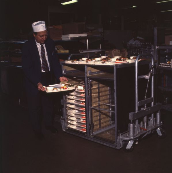 A man wearing a white chef cap and a suit and tie, is either placing trays of food on a cart, or taking them off for a Pan American World Airways Boeing 747 airplane.