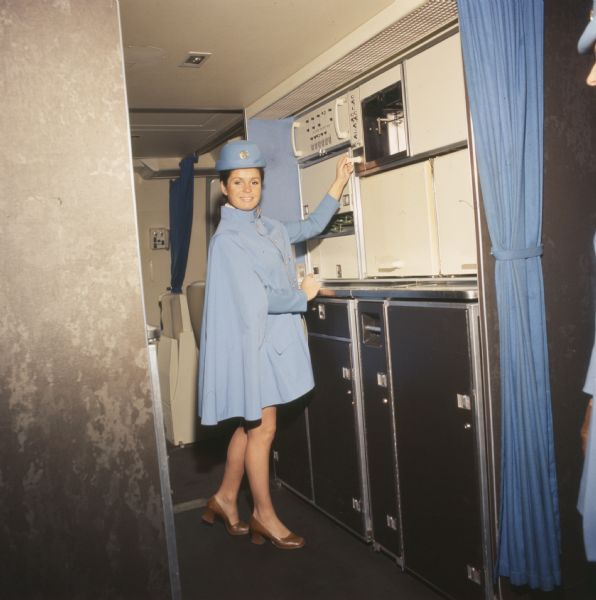 A female flight attendant in uniform is standing in a Pan American World Airways Boeing 747 airplane. She is standing in front of a set of cabinets, and is holding onto a switch near controls in front of her.