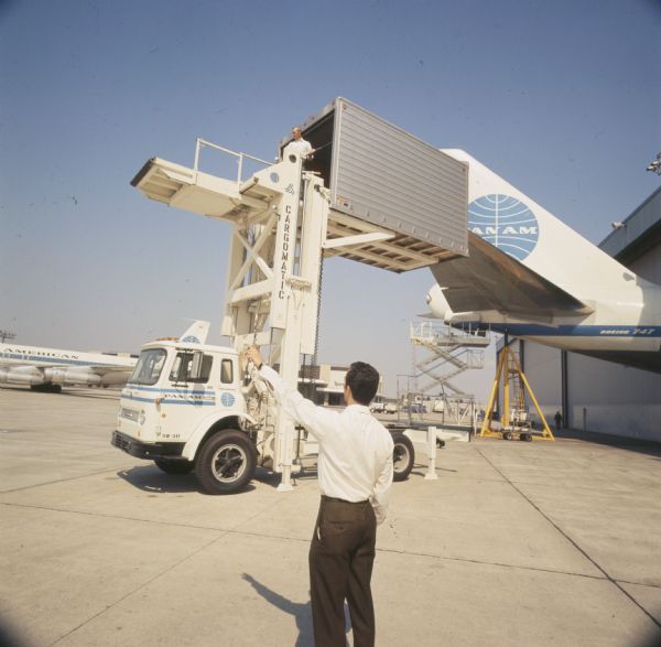 Color photo of an International truck carrying prepacked airline food to Pan American World Airways Boeing 747 airplane. A man in the foreground gestures to his left while an International truck with a lift platform called a "Cargomatic" is raising a man and cargo container into the air. The photo caption reads: "Representative sampling of this service armada... CO-Loadstar truck, a galley servicing van. It carries to plane modules, prepacked with finest cuisine, elaborate place settings."