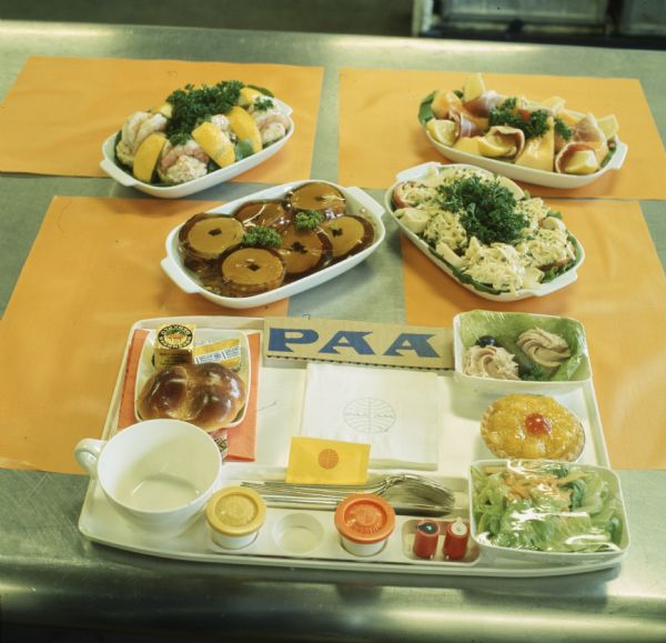 Food tray for a Pan American World Airways Boeing 747 in-flight meal. The tray has silverware, a coffee cup, and assorted food items. There is also a napkin with the Pan Am logo. Four entrees are arranged above the tray.