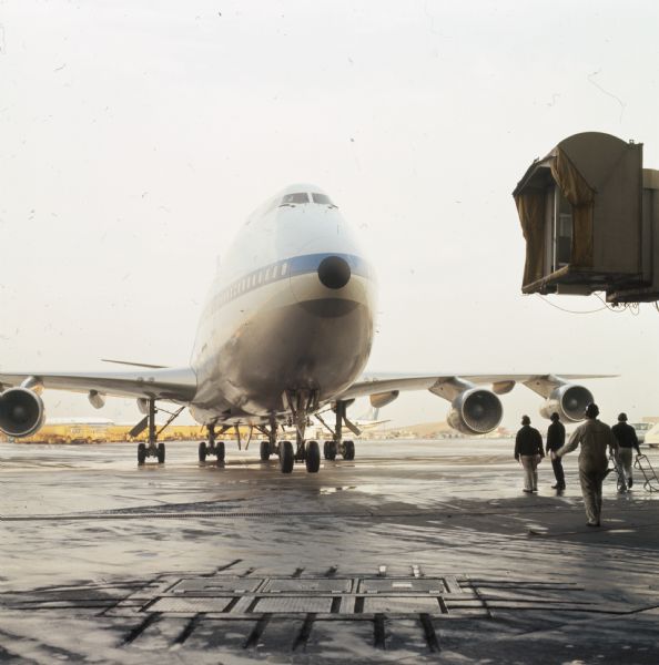 A Pan American World Airways Boeing 747, with what may be a ground crew walking towards it. A passenger boarding bridge is suspended in the air on the right.