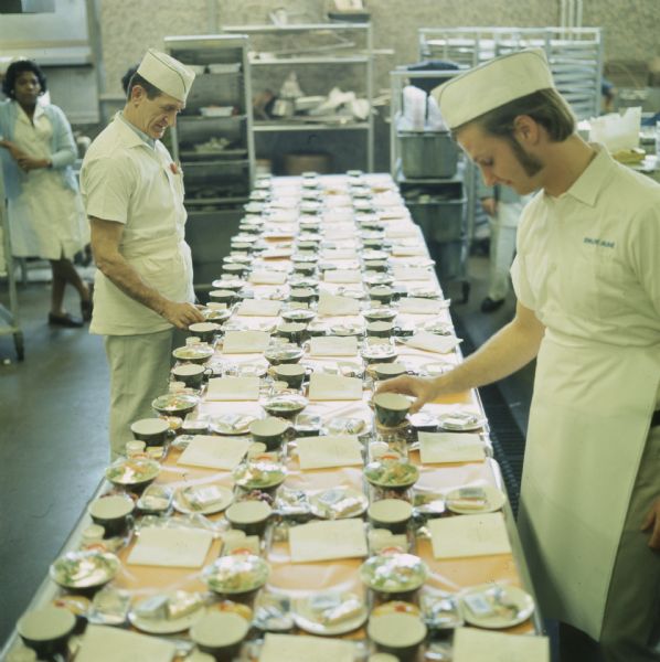 Two male food service employees preparing trays of food laid out on a table for a Pan American World Airways Boeing 747 in-flight meal. A woman is standing in the background on the left. Shelves and trays are along the back wall.