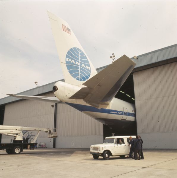 Color photo of the tail end of a Pan American World Airways Boeing 747 sticking out of a maintenance shed. In the foreground is an International Scout, possibly a model 800-A. One man sits in the Scout, while talking to three men. Another vehicle with a lift bucket attached to the back is in the background on the right.