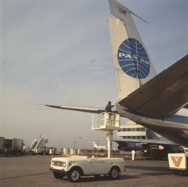 Color photo of an International Scout with a man in a lift platform raised up to the tail of a Pan American World Airways Boeing 747 parked at an airport.