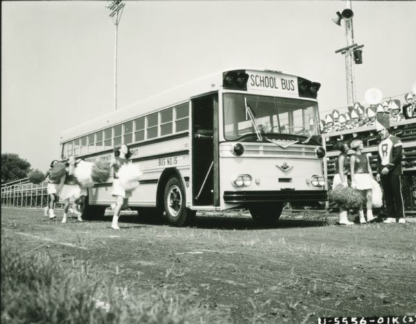 Cheerleaders surround an International school bus with Wayne body parked on a football field. A band is sitting in the bleachers behind the bus.