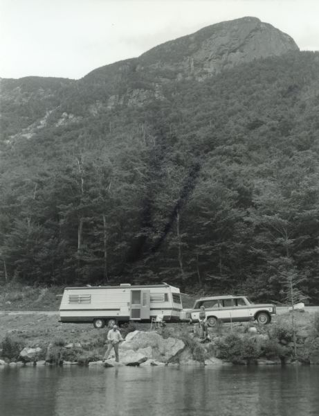 An International Scout hauling a camper is parked next to a body of water. A man is fishing while a woman looks on from the banks of the water.