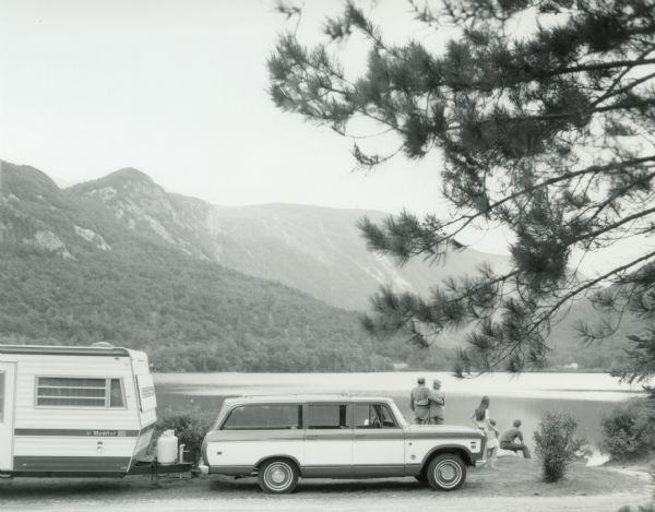 View towards lake and mountain with an International Scout hauling a camper parked next to the shoreline. A man, woman, and three children are gathered behind the Scout looking out at the lake.
