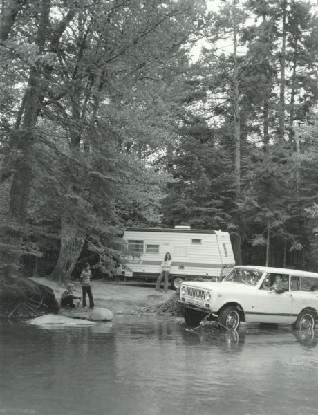 View across water of a man in the driver's seat of an International Scout parked in the water at the shoreline of a lake. On the left posing at the shoreline is a young boy holding a fishing pole, and a young girl standing. Behind them a woman is standing and leaning on the camping trailer in the background.