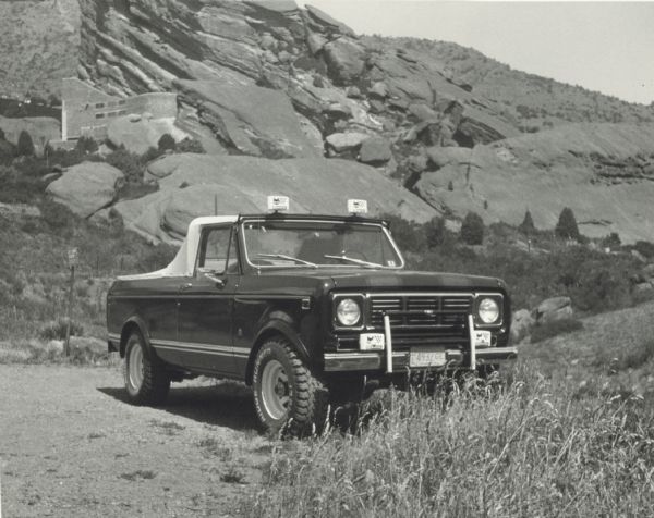 An International Scout "Suntanner" parked in the foreground, with a large rock formation in the far background, somewhere in the southwest United States. Halfway up the formation is a stone wall or building.