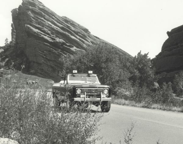 A woman drives an International Scout "Suntanner" on a highway in the Southwest United States. In the background are large rock formations and vegetation.