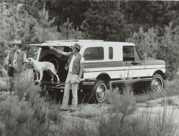 An International Scout Terra pickup with cap is parked in a wooded area with two men standing near a dog standing on the open tailgate. The men are holding guns, presumably for a hunting trip.