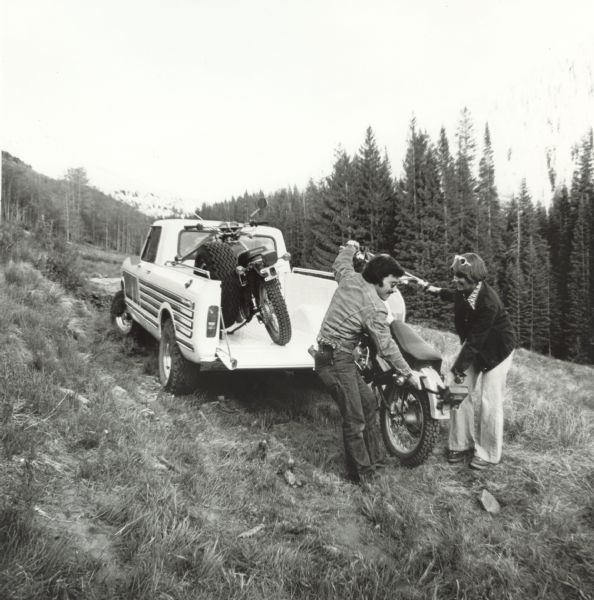 A man and woman load a motorcycle into an International Scout Terra parked on a hill in a wooded area.