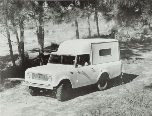 A man in the driver's seat of an International Scout 80 with Camper parked in front of trees.