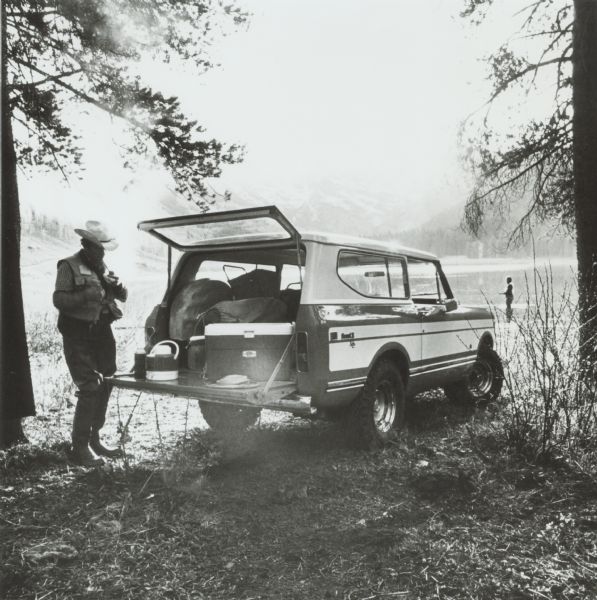 An International Scout II XLC parked on the shoreline of a lake. A man is using the hatchback of the truck to prepare for fishing, while another man in the background is standing in the lake fishing.