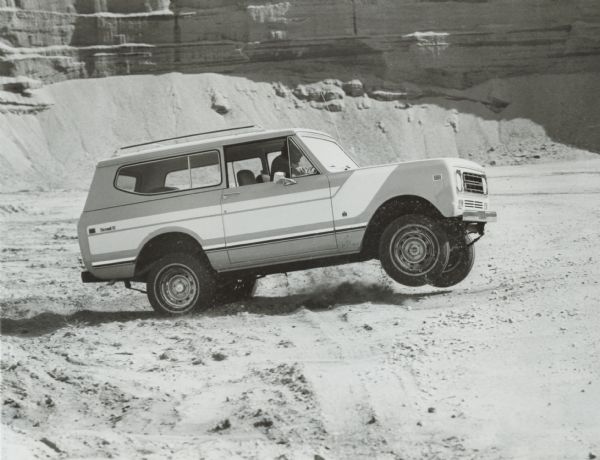 Side view of a man driving an International Scout II in the desert. Rock formations are in the background. Caption with photograph reads: "International Scout II Four-wheel-drive sport utility compact for 1977 features a restyled grille insert, two new exterior colors--"Siam Yellow" and "Elk" and a new "Parchment" vinyl interior. The Scout is the most popular model ever produced by International Harvester's Truck Division--almost 400,000 have been sold since the vehicle was introduced in 1961."