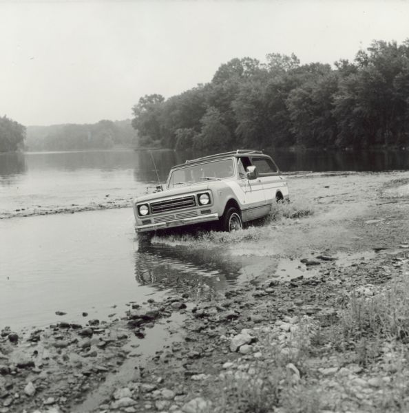 An International Scout driven off-road through a body of water along a shoreline, probably a river or lake.