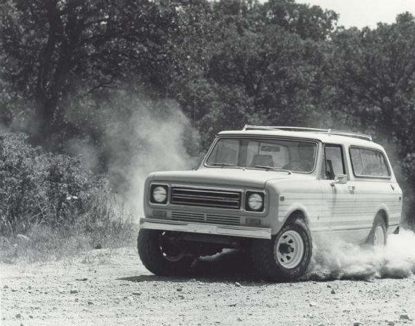 A man is driving an International Scout Traveler on a dirt road. The wheels are creating clouds of dust.