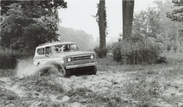 A person is driving an International Scout II off-road in a sandy area among trees. The spinning wheels are churning up clouds of dust.