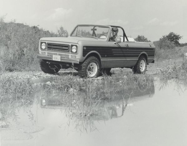 A man is sitting in the driver's seat of an International Scout Terra which is parked at the edge of a lake or pond. Photograph was taken for International Harvester by Brandt & Assoc., Ltd. Photography of Barrington Hills, Illinois.
