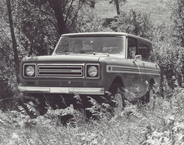 Two men are riding in an International Scout which is pictured off-roading in a wooded area.