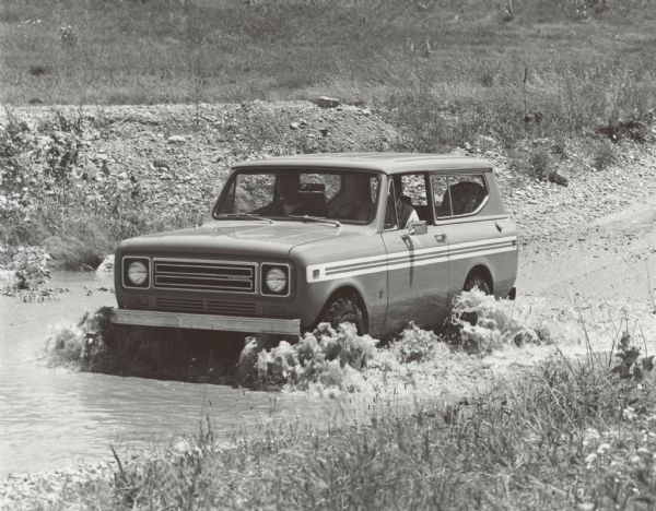 Two men are in an International Scout which they are driving from a gravel road into the water.